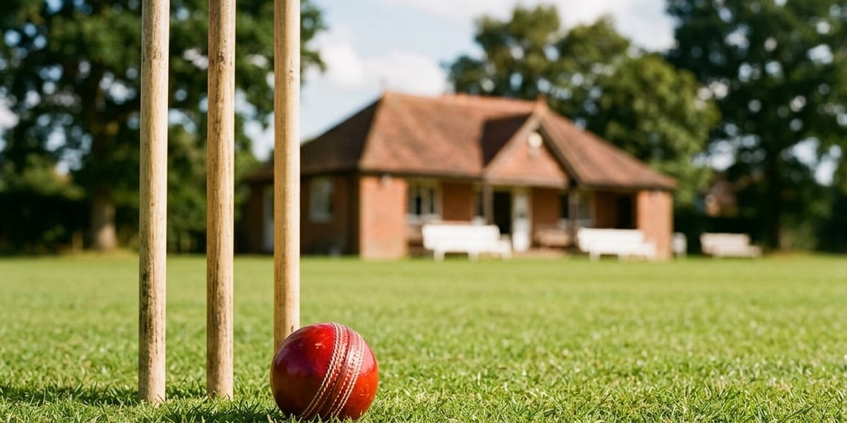 Red cricket ball resting on the grass beside a set of wooden stumps, with a brick pavilion behind on a sunny summer afternoon